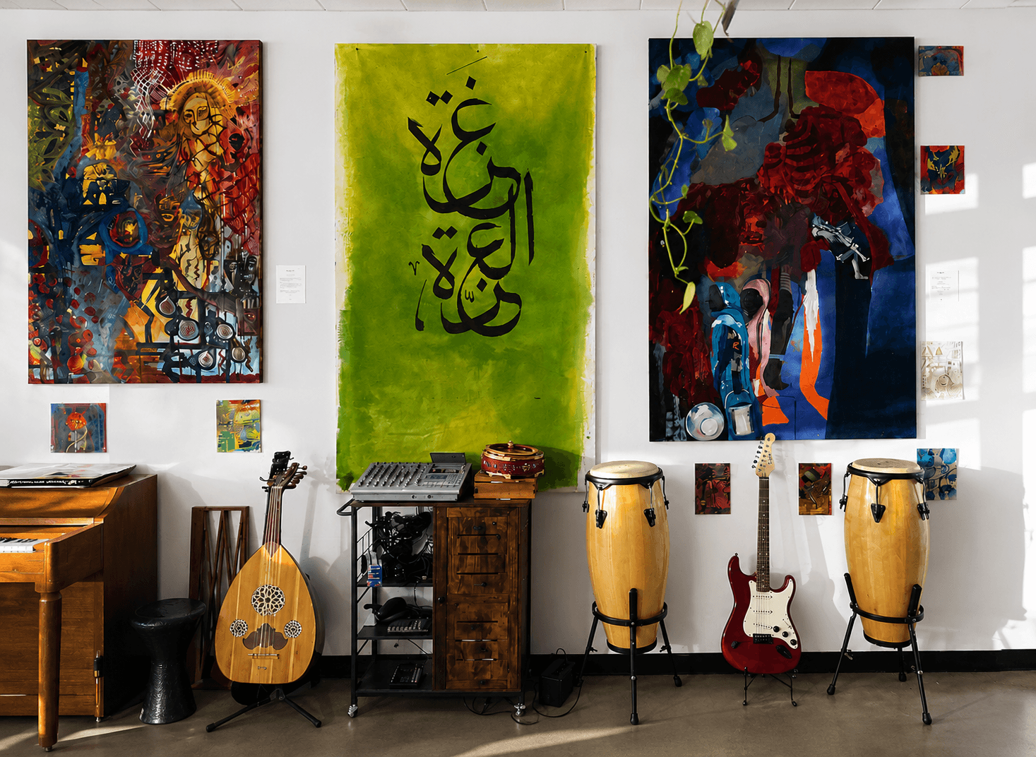 A sunlit studio wall at the Jerusalem Academy hung with three large paintings — including a lime-green canvas with bold black Arabic calligraphy reading Gaza — above an upright piano, a wooden oud, an electric guitar, congas, and audio equipment
