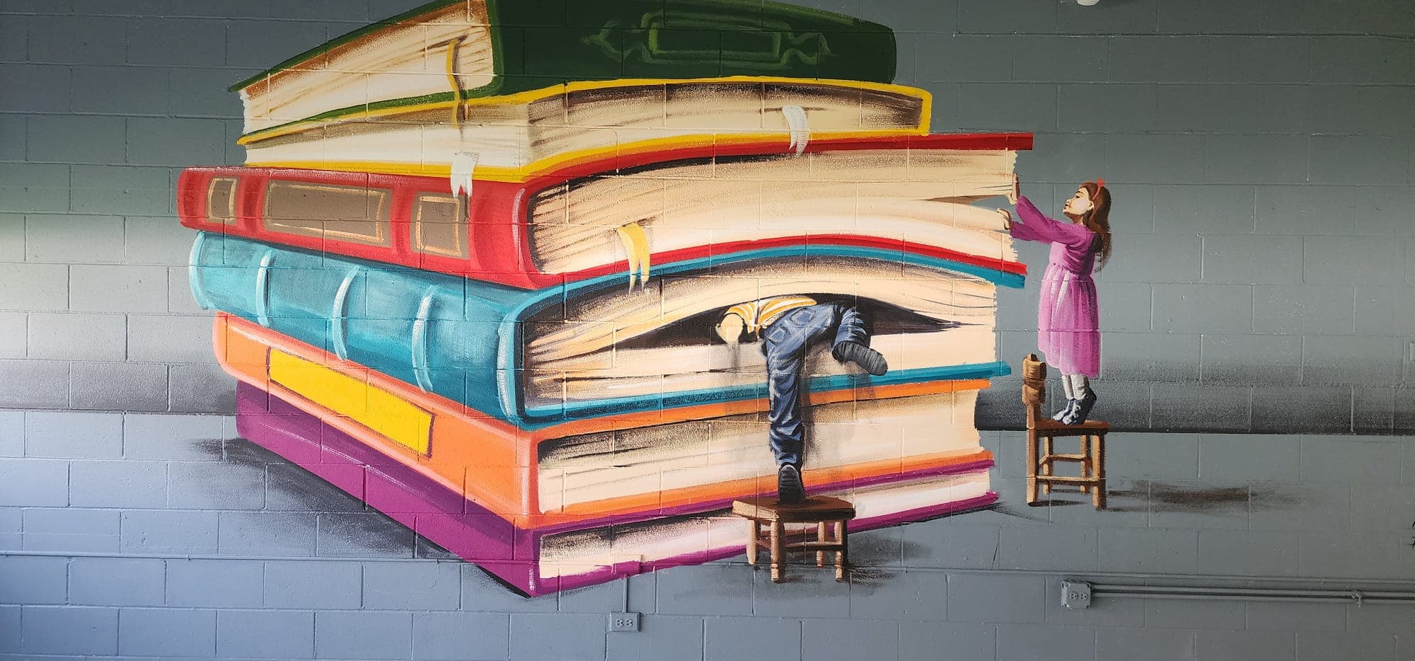 Interior mural of a giant colorful stack of books, with children on wooden stools climbing up and peeking out from between the pages, on a gray cinderblock wall.