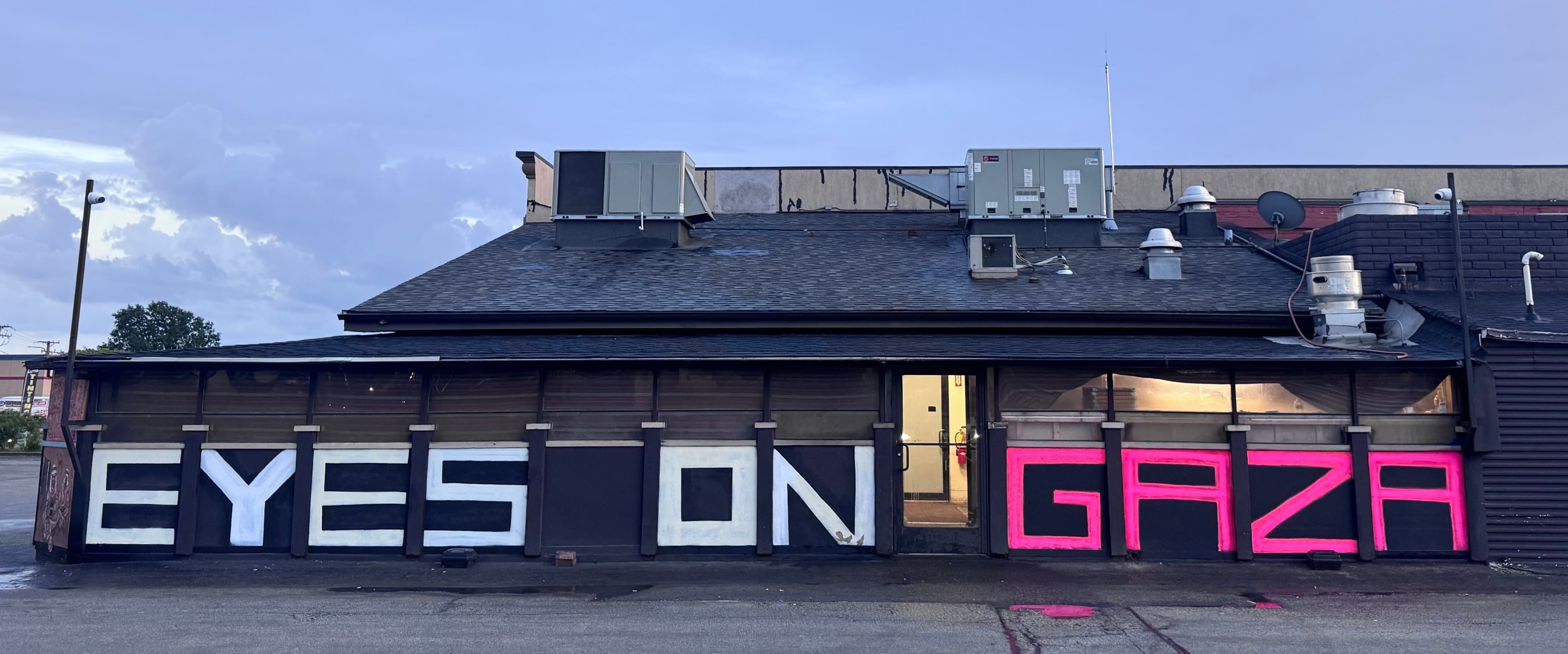 A storefront building at dusk with "EYES ON GAZA" painted in large white and hot-pink block letters across its black lower facade.