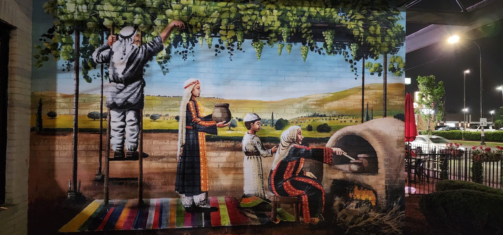 Wall mural of a Palestinian countryside scene — a man harvesting grapes from a vine trellis, a woman in an embroidered thobe carrying a clay jug, a child, and an elder baking bread at a taboon oven, over golden hills and a woven rug.