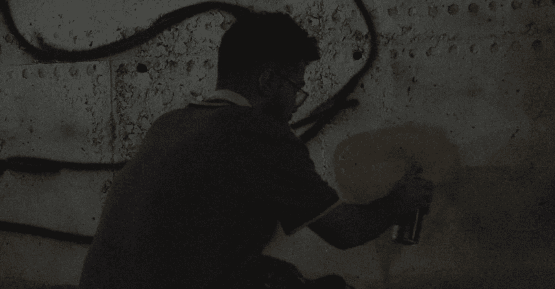 Film still: an artist crouched spray-painting graffiti on a concrete wall in low light