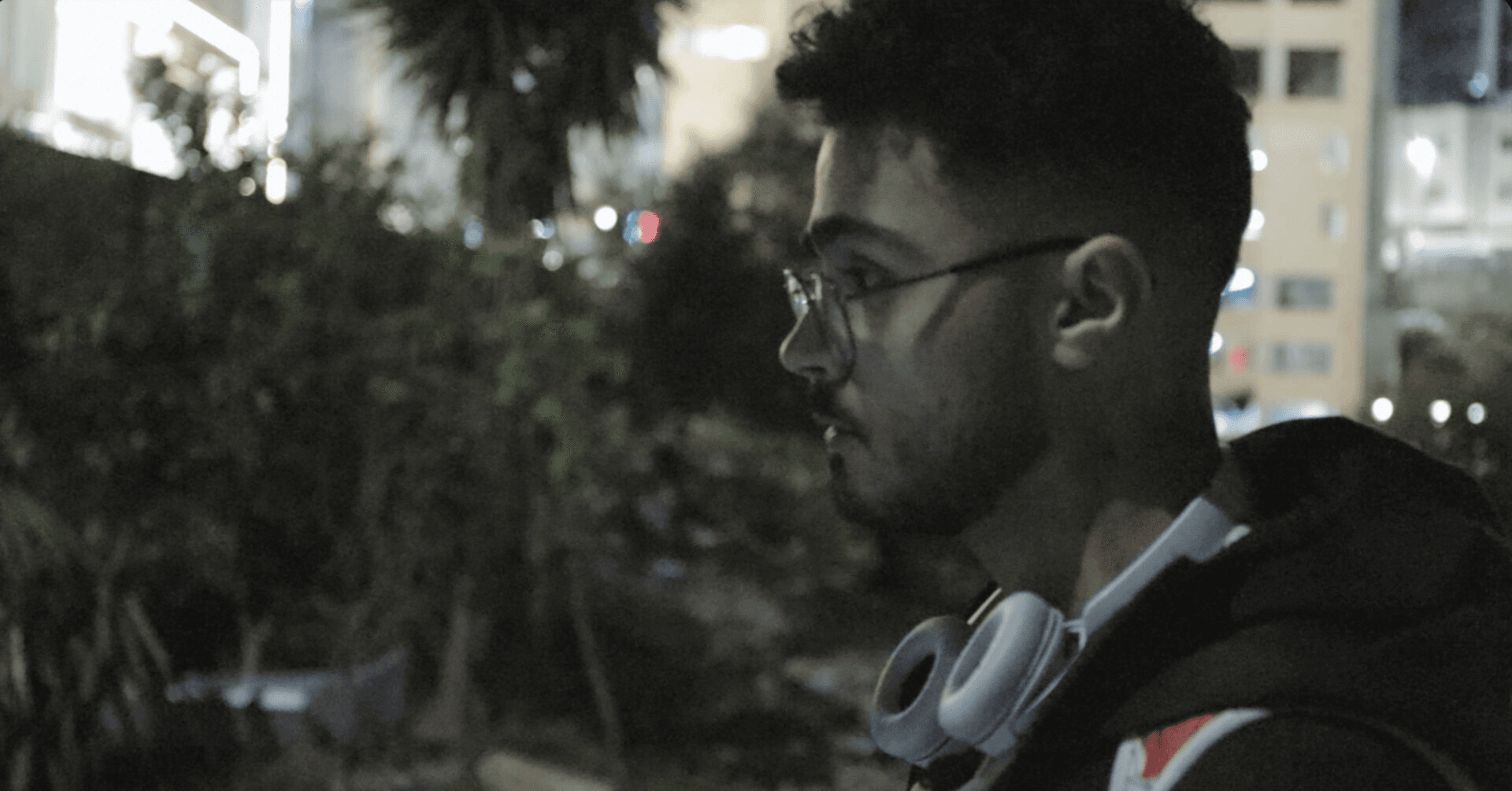 Film still: a night profile portrait of a young man in glasses with headphones around his neck