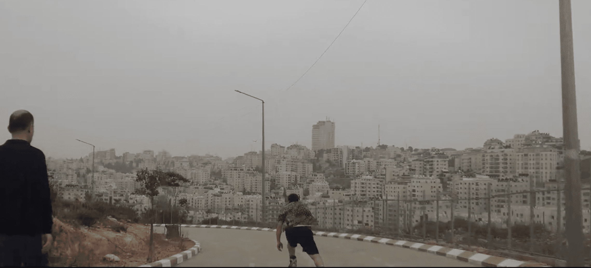 Film still: two people skating down an empty road overlooking the hillside cityscape of Ramallah