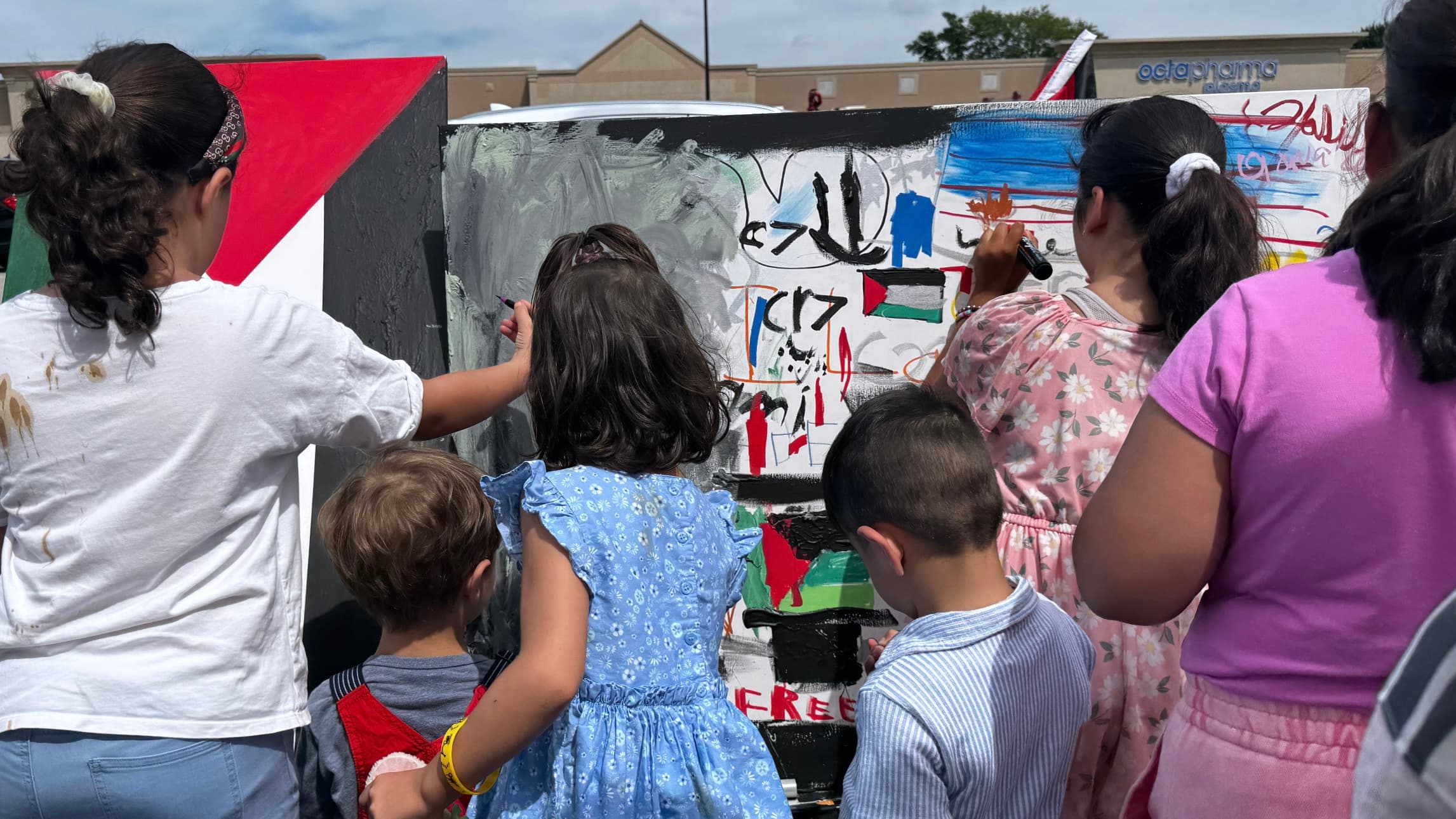 Children and youth painting a board in Palestinian flag colors and Arabic script at an outdoor rally
