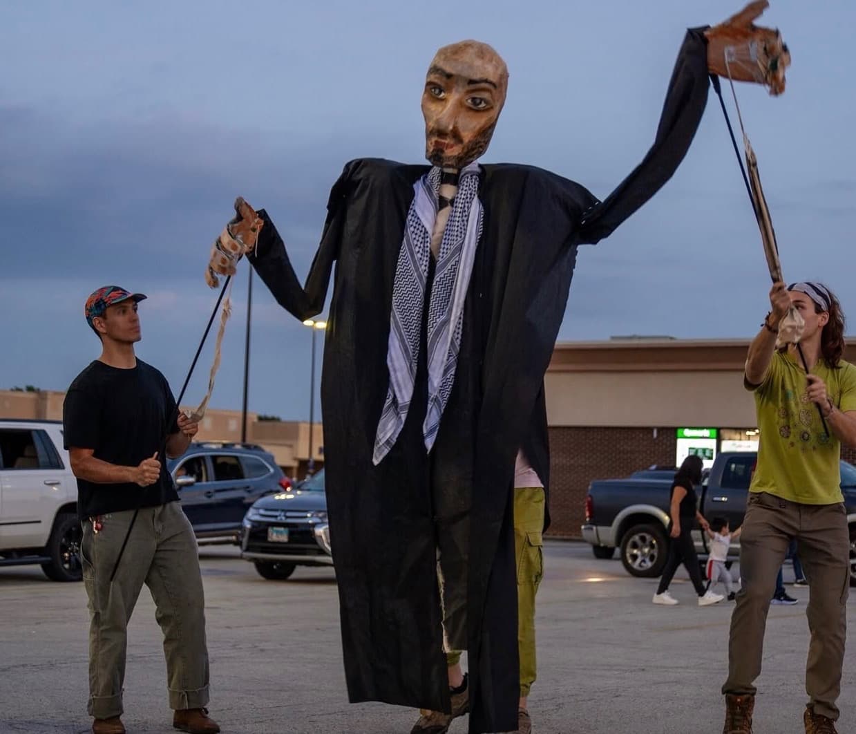 A tall papier-mache puppet in a black robe and keffiyeh worked by two puppeteers in a parking lot