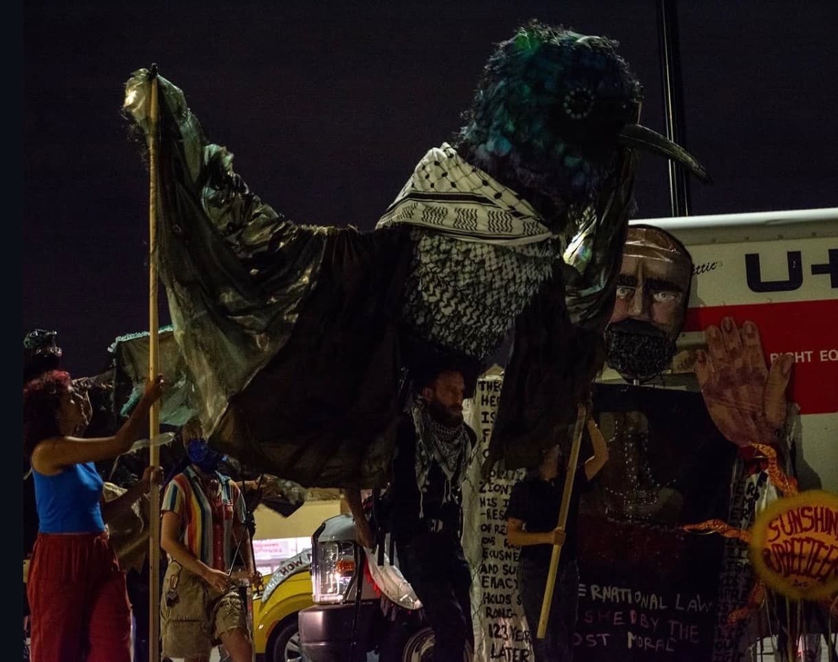 A giant iridescent sunbird puppet draped in keffiyeh cloth, raised on poles at night