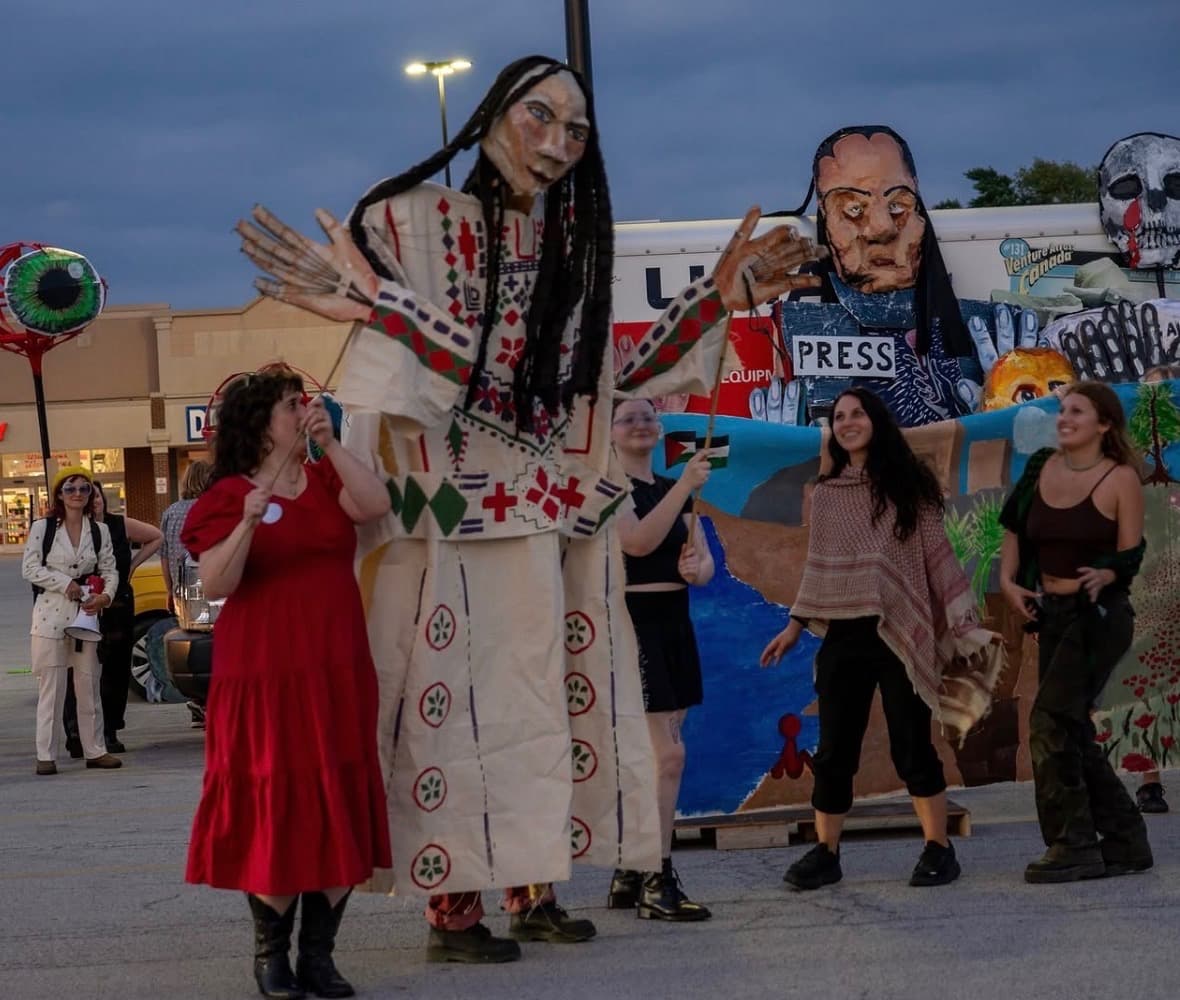 A giant puppet in an embroidered Palestinian thobe towering over dancers at dusk during the parade